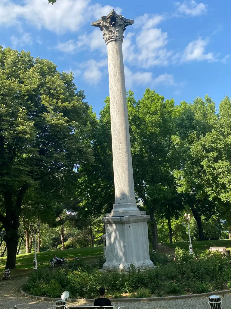 Gotlar Sütunu; kaidenin dibinde çiçek tarhı, arkada yeşil ağaçlar, önde parke yol ve beyaz bulutlu mavi gök.