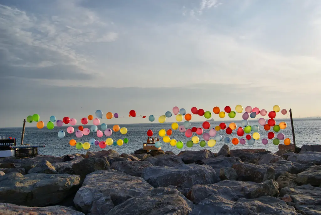 Moda sahilinde kayaların üstünde renkli balonlar; gerili ipte dizilmiş balonlar, sakin deniz ve bulutlu gökyüzü.