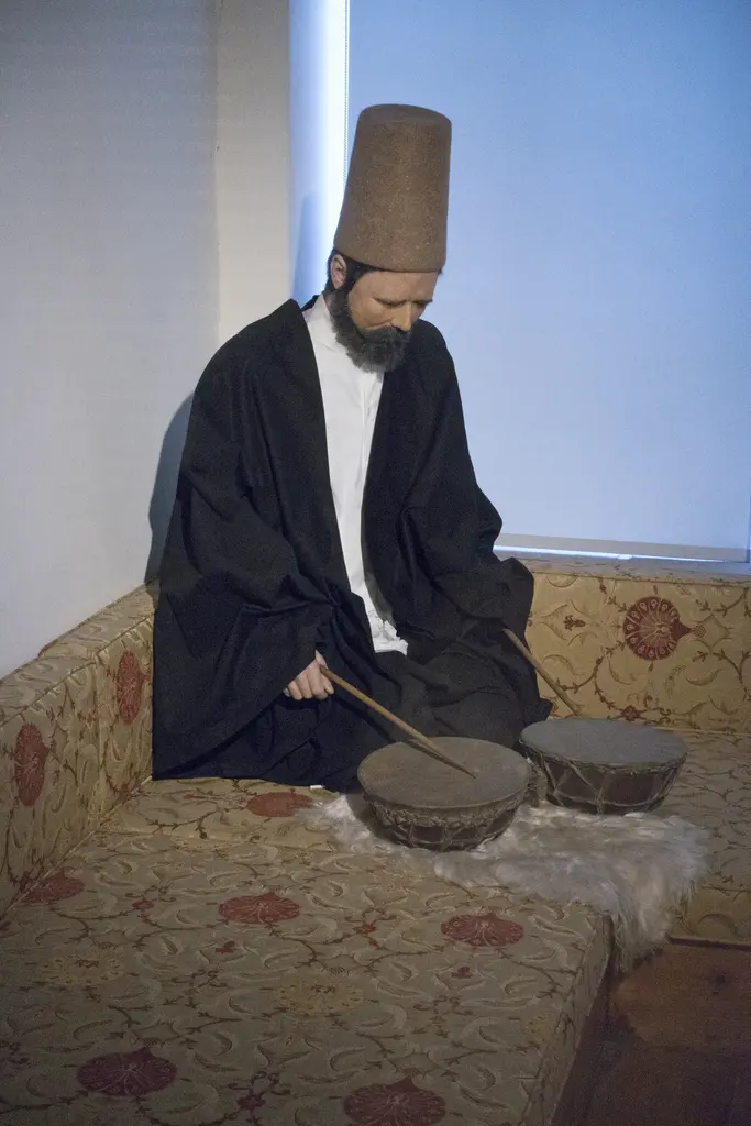 Müzede sikkeli ve siyah hırkalı bir derviş mankeni; minderde oturmuş, önündeki kudüme vurur hâlde.