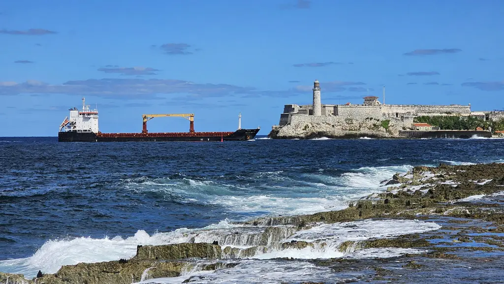 Havana limanının girişinde El Morro Kalesi; kayalık burnun üzerindeki taş surlar, yanındaki beyaz deniz feneri, önünden geçen yük gemisi ve kayalıklarda kırılan dalgalar.