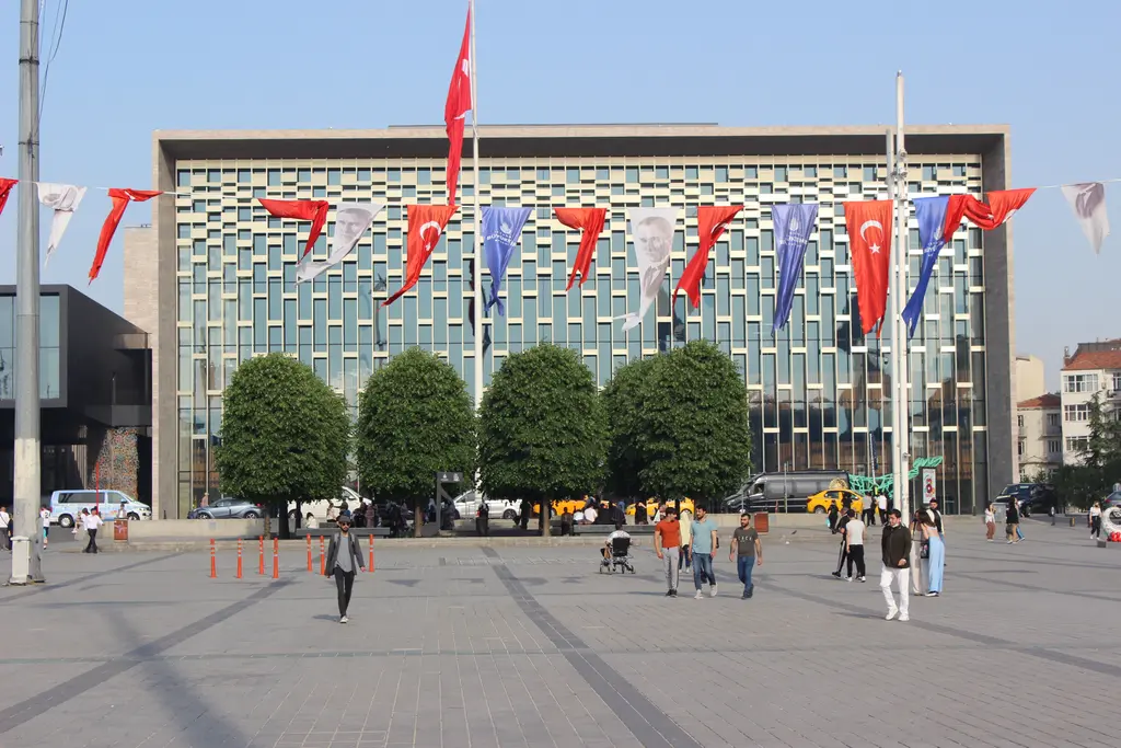 Atatürk Kültür Merkezi'nin önü; budanmış yuvarlak ağaçlar, bayrak dizileri, meydanda yürüyen insanlar ve arkada cam cephe.