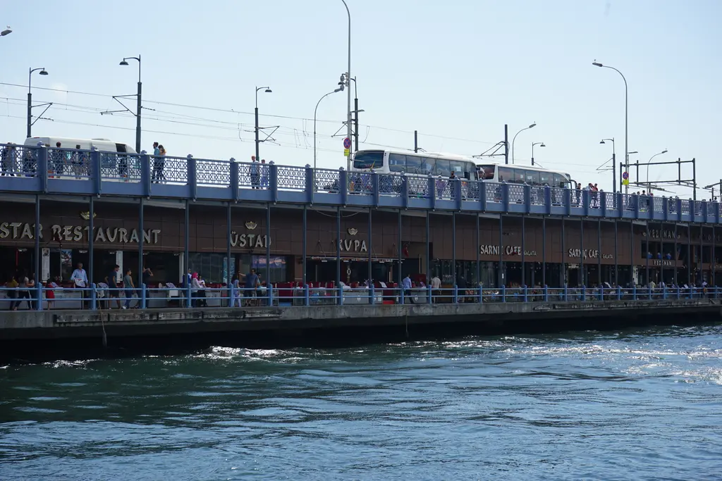 Galata Köprüsü'nün alt katı; korkuluk boyunca lokanta tabelaları, üst katta yürüyen insanlar ve önde dalgalı su.