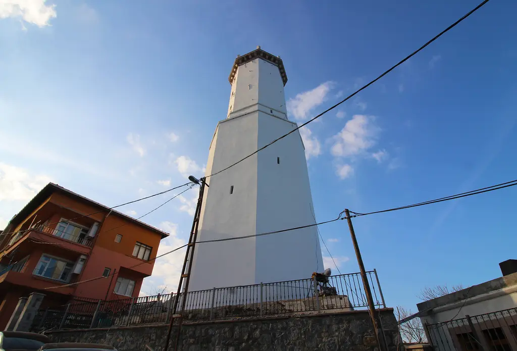 Rumelifeneri'nde fener; mavi gökyüzüne yükselen beyaz kule, yanında turuncu cepheli bir ev ve elektrik telleri.