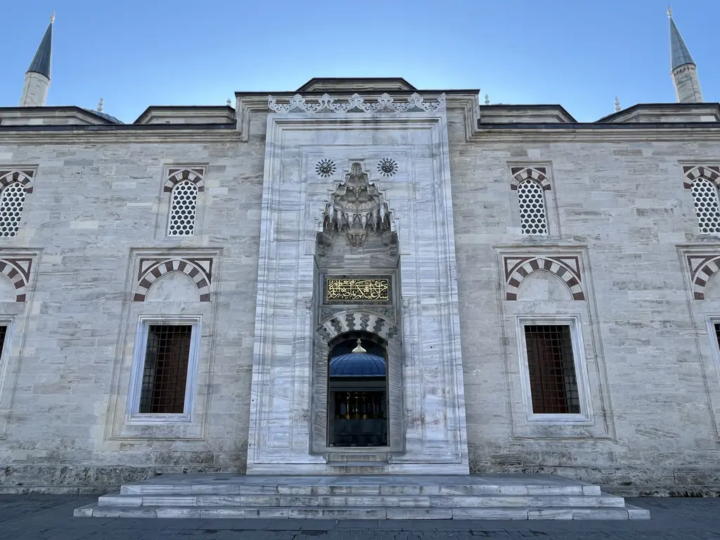 Avlunun mermer taç kapısı dışarıdan; mukarnaslı kavsara, altın yaldızlı kitabe levhası, iki yanda şebekeli pencereler ve arkada iki minare.