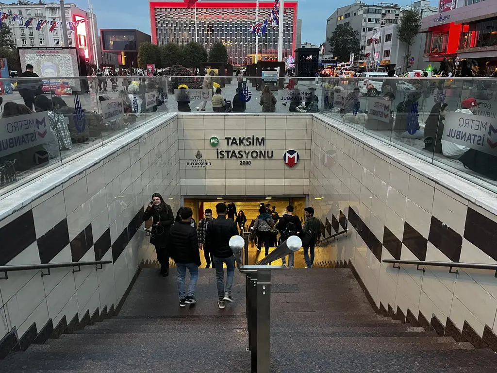 Taksim'de M2 metrosunun meydana açılan girişi; merdivenlerden inen insanlar, Taksim İstasyonu tabelası ve arkada AKM'nin cephesi.