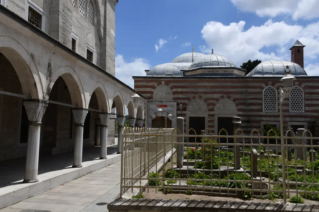 Mihrimah Sultan Camii'nin haziresi; demir parmaklıklı alan içinde sarıklı mezar taşları, arkada caminin yan revakı ve kubbeli almaşık yapı.