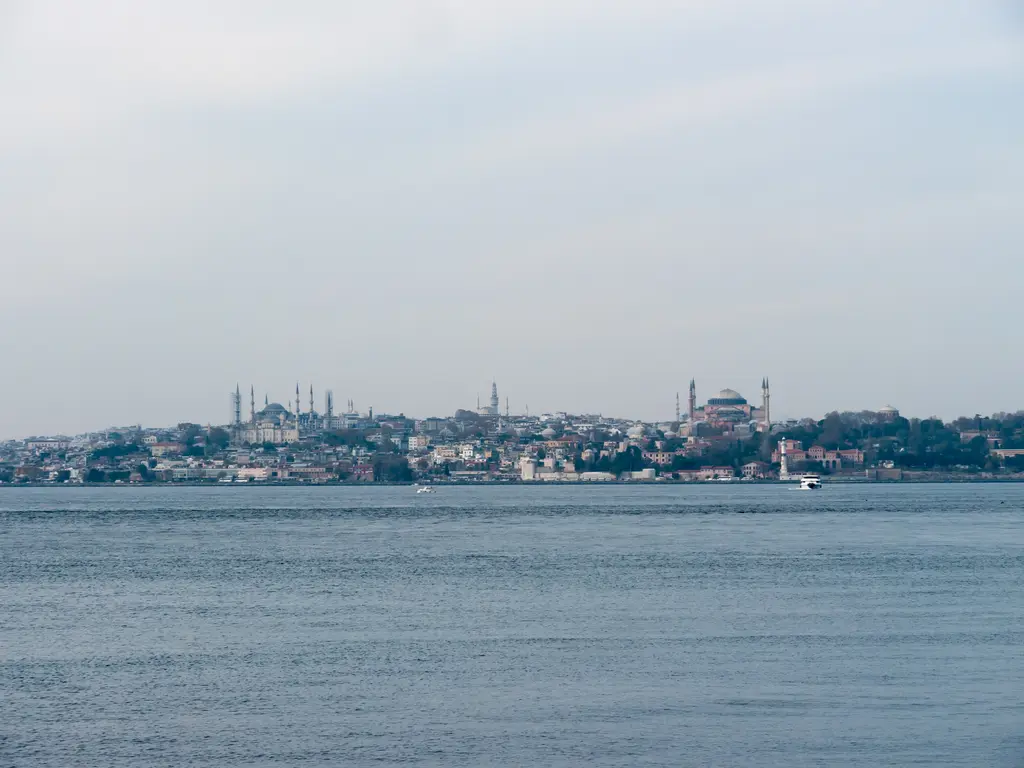 Moda kıyısından tarihi yarımada; sakin gri deniz, ufukta tarihi yarımadanın kubbeleri ve minareleri, puslu gökyüzü.