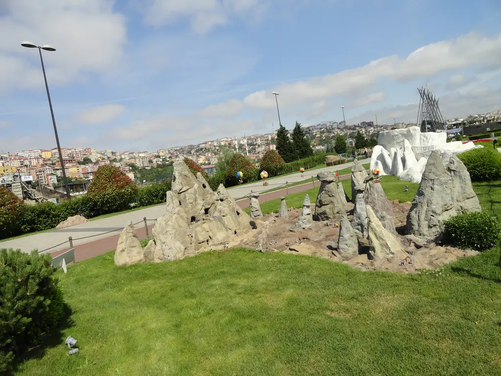 Miniatürk'te Kapadokya maketi; güneşli çimen üzerinde gri peri bacaları, arkada beyaz Pamukkale maketi, yollar ve tepedeki evler.