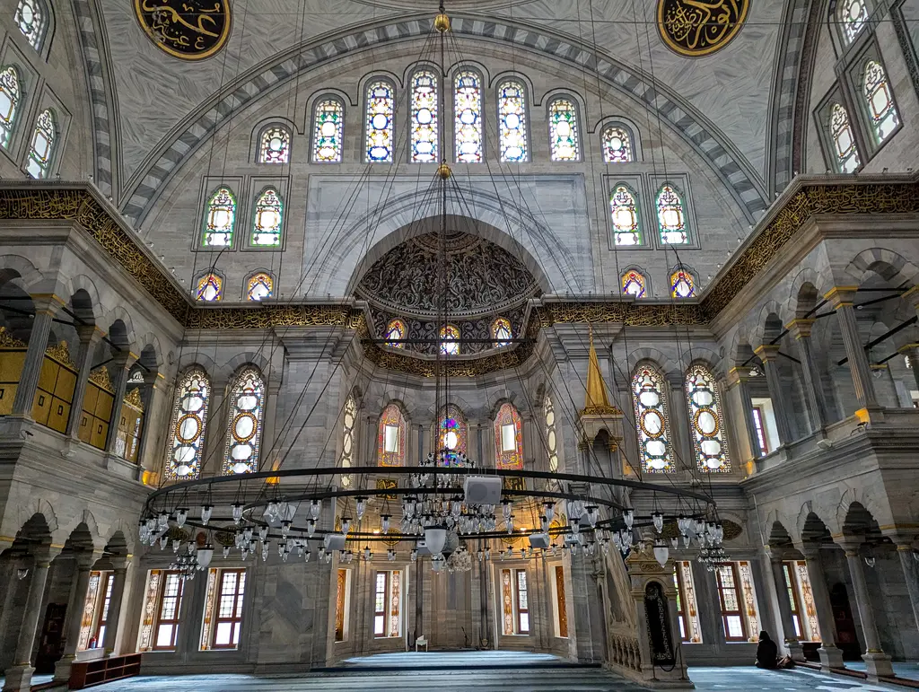 Nuruosmaniye Camii'nin harimi mihrap yönüne bakış; sıra sıra renkli cam pencereler, halka biçimli avize, sağda minberin külahı ve sıcak sarı ışık.