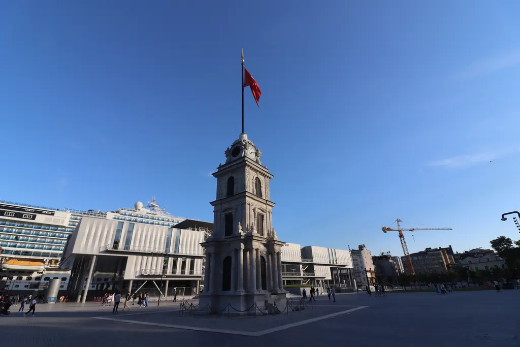 Tophane'de geniş taş meydanda saat kulesi; tepesinde Türk bayrağı, arkasında Galataport'un alçak binaları.