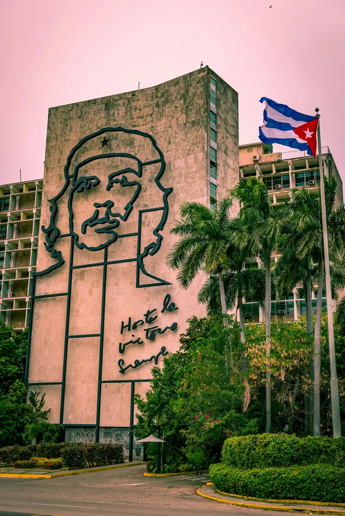 Havana'da bir bakanlık binasının cephesindeki çelik hatlarla yapılmış Che Guevara portresi; yanında dalgalanan Küba bayrağı, önünde palmiyeler ve bakımlı yeşillik.