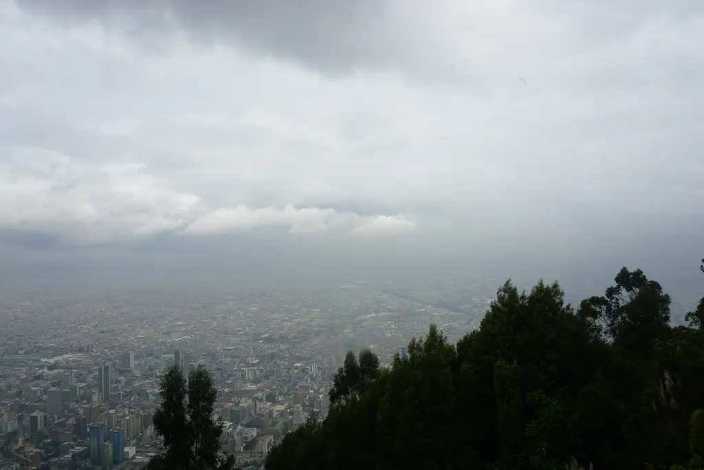 Bogotá'da şehir manzarası; yüksekten bakıldığında ufka kadar uzanan yoğun kent dokusu, önde ormanlık yamaç ve üzerinde alçak bulutlar.
