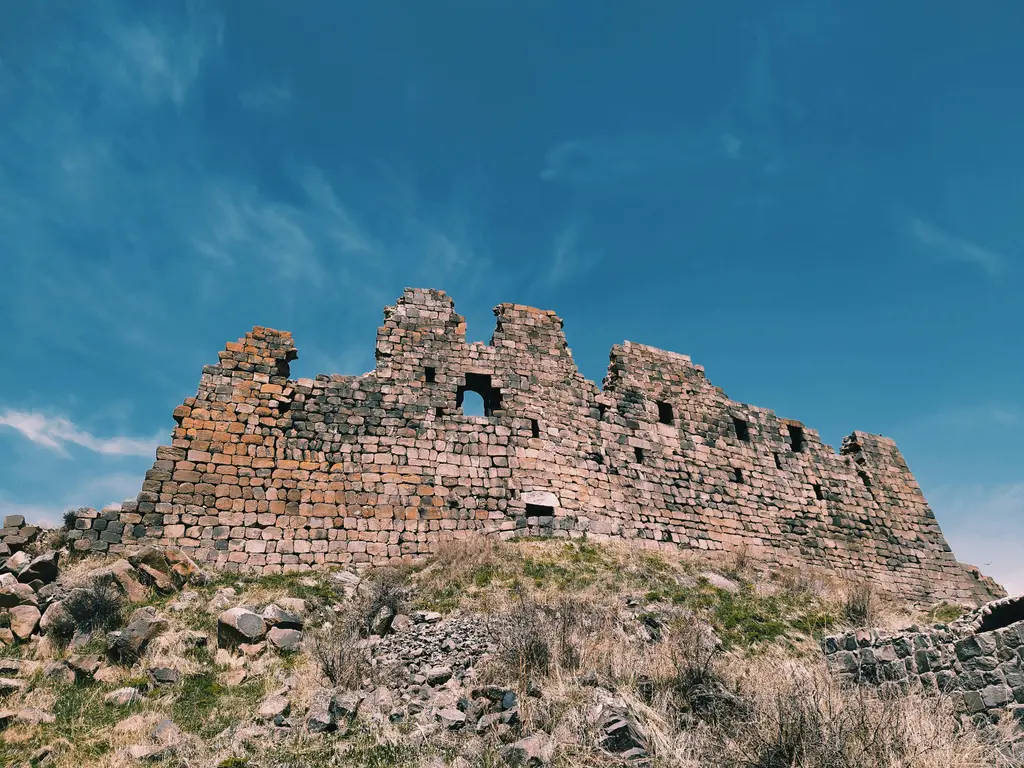 Aragats yamacında sarı ve gri taş bloklardan örülmüş, üstü yıkılmış kale duvarı; duvarda küçük bir pencere boşluğu, önde kuru otlar ve dağılmış taşlar, üstte koyu mavi gökyüzü.