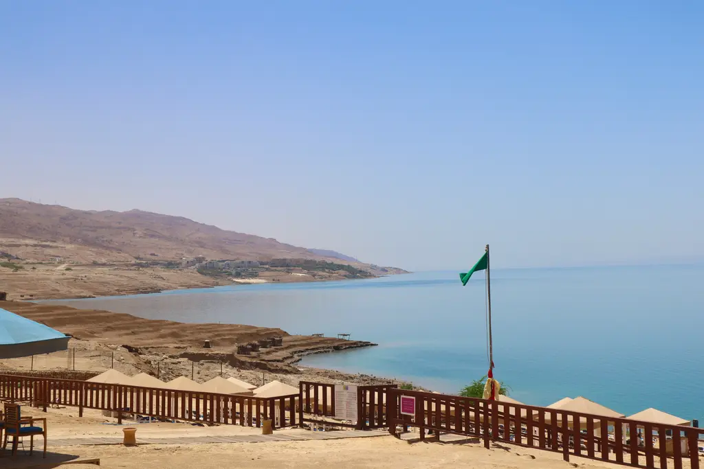 Ölü Deniz kıyısı; ahşap korkuluklu bir teras, direkte dalgalanan Ürdün bayrağı, kıyıya vuran açık renkli su ve karşı kıyıdaki kurak dağ silsilesi.