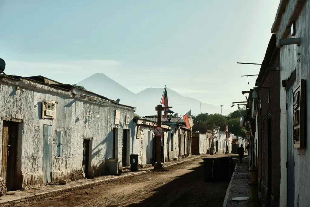 San Pedro de Atacama; kerpiç duvarlar arasındaki tozlu sokak ve alçalan güneşte uzakta görünen yanardağ konisi.