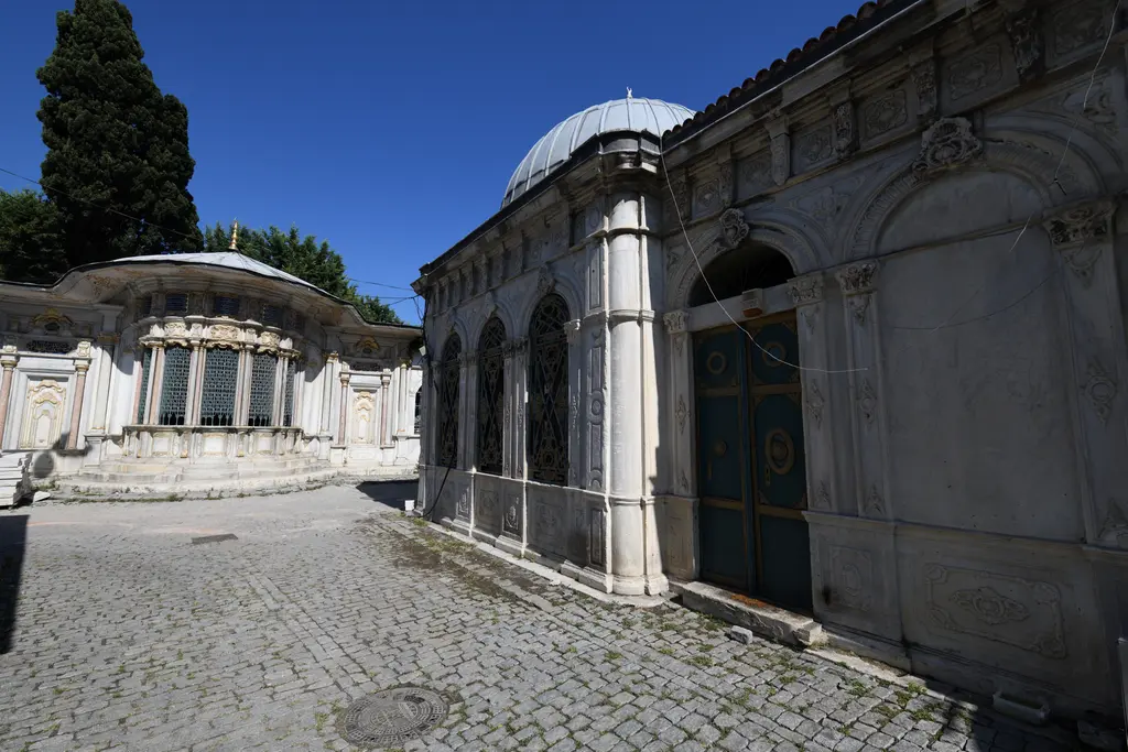 Mihrişah Valide Sultan Sebili ile türbe; dalgalı saçaklı mermer sebil, yanında türbenin şebekeli pencereli duvarı ve taş döşeli sokak.