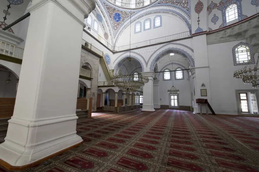 Gazi Atik Ali Paşa Camii'nde kalın beyaz payeler; kemerlerin ardında yan bölme, kalem işli kubbeler, pencereler ve desenli halı.