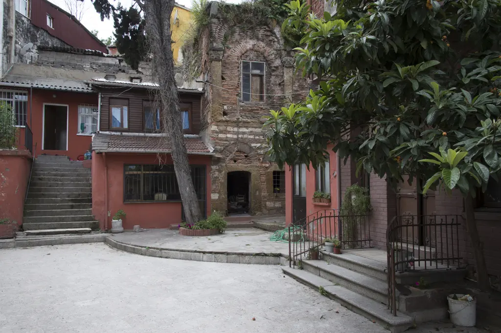 Panayia Balino Kilisesi avlusu; taş döşeli geniş avlu, merdivenle çıkılan kırmızı boyalı bina, ağaçlar ve arkada tuğla cepheli yapı.