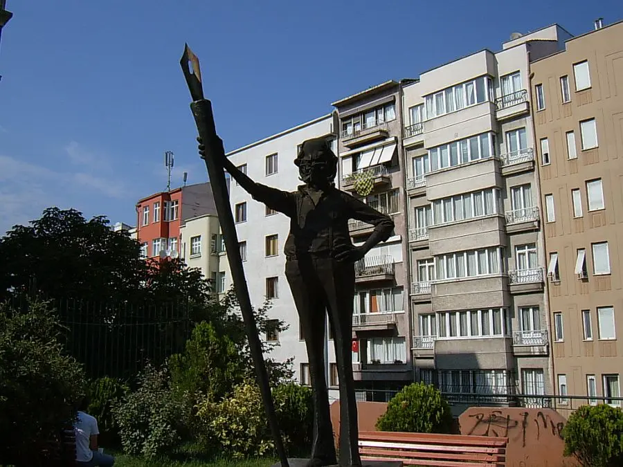 Cihangir Parkı'nda Oğuz Aral heykeli; elinde dev bir kalem tutan bronz figür, arkada apartmanlar ve ağaçlar.