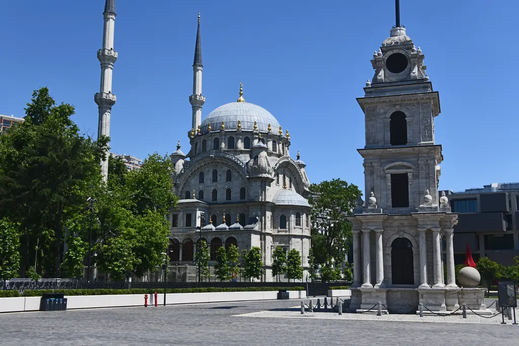Tophane'de taş döşeli meydan; ortada dört katlı saat kulesi, arkasında iki ince minareli kubbeli cami ve yeşil ağaçlar.