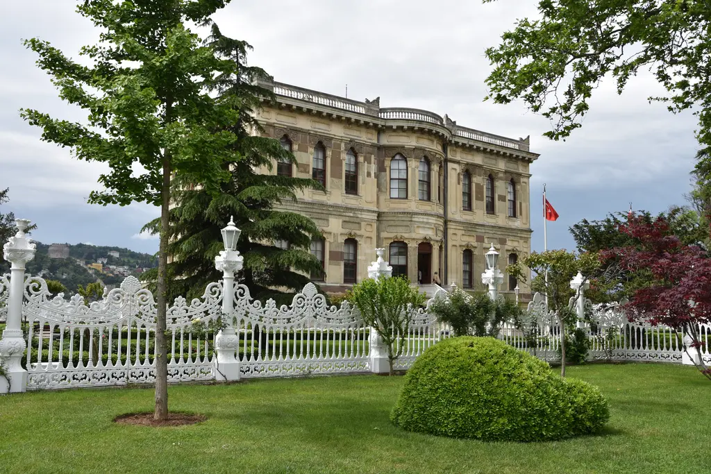 Küçüksu Kasrı; bahçede süslü cepheli taş kasır, oymalı beyaz parmaklıklar, çimenlik ve genç bir ağaç.