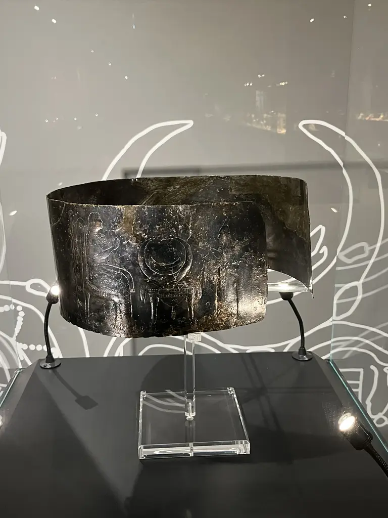 Ziyafet Sahneli Geniş Kemer'e yakından bakış; cam kaide üstünde yay biçimli bronz levha, üzerinde işlenmiş figürler ve koyu gri fon.