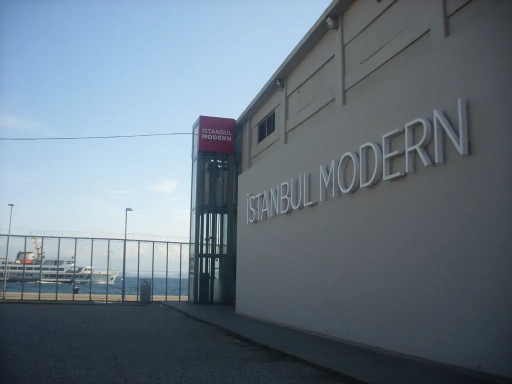 Eski İstanbul Modern binasının gri duvarı; büyük "İSTANBUL MODERN" harfleri, kırmızı asansör kulesi ve arkada Boğaz.