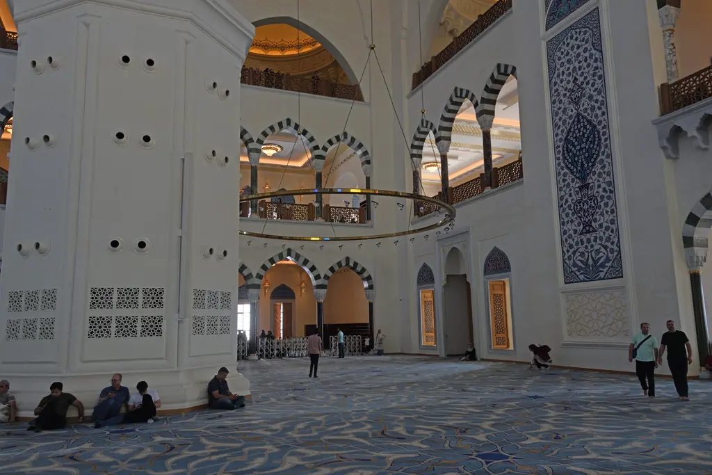Büyük Çamlıca Camii'nin içi; beyaz payeler, kemerli mahfil katları, desenli halı ve oturan ziyaretçiler.