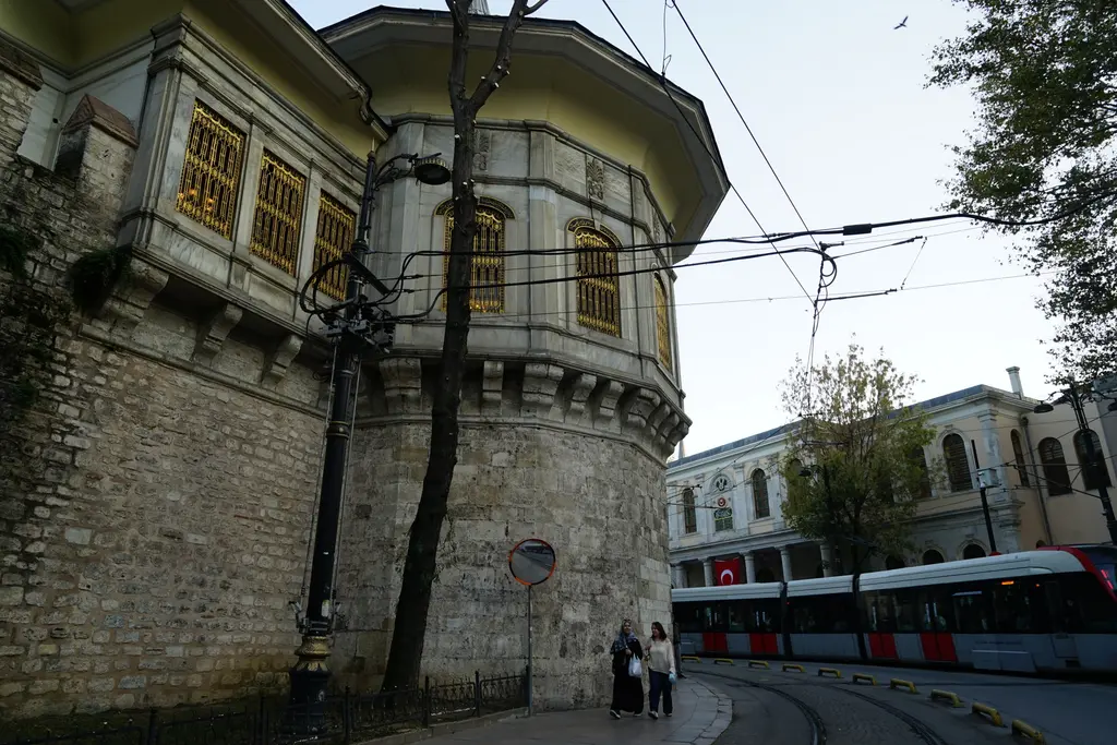 Alay Köşkü'ne alttan bakış; taş burç ve sur, üstte kafesli pencereli salon ile kanat, sağda geçen tramvay ve karşı binalar.