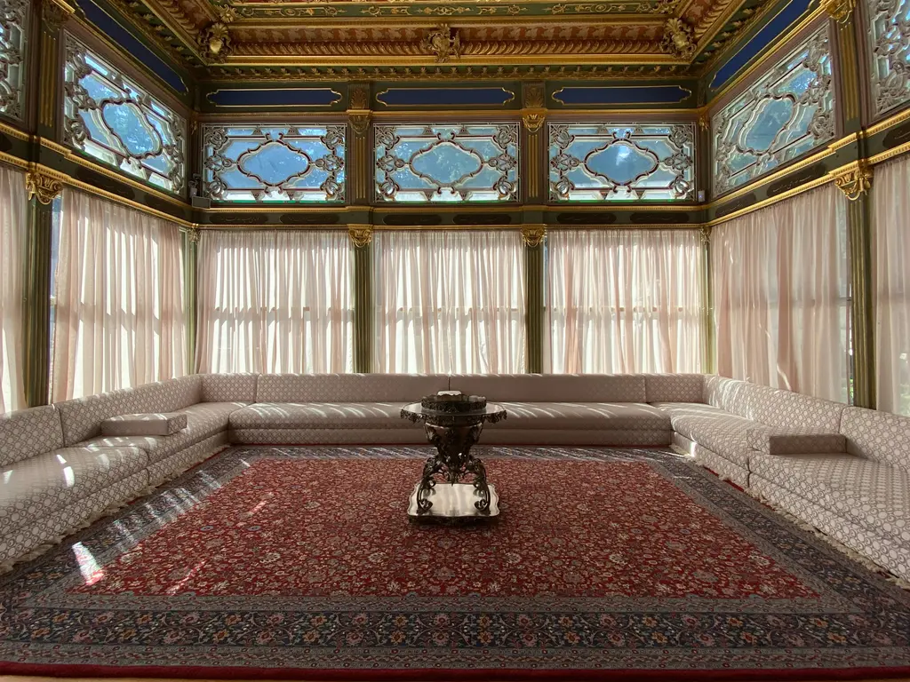 Topkapı Sarayı'nda bir köşkün iç mekânı; üç yanı saran alçak sedir, kırmızı desenli halı, renkli camlı pencereler ve altın işlemeli ahşap tavan.