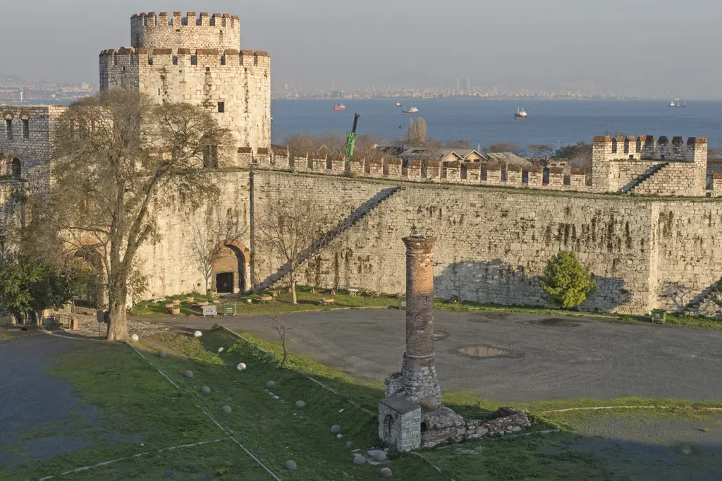 Yedikule Hisarı avlusunda kaide üzerinde duran tek yivli mermer sütun; arkada kemerli girişi olan yuvarlak kule, yapraksız ağaçlar ve uzakta Marmara Denizi.