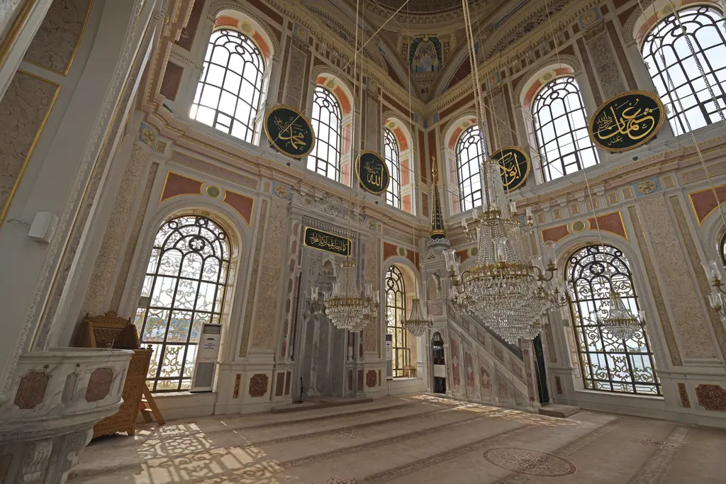 Ortaköy Camii'nde harim; pencerelerden giren ışık, sağda mermer vaaz kürsüsü, avizeler, hat levhaları ve halı.