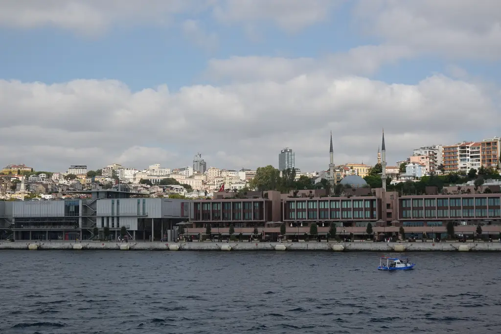 Denizden Galataport; önde dalgalı su, kıyıda alçak tuğla rengi binalar, arkada Tophane yamacı, minareler ve apartmanlar.
