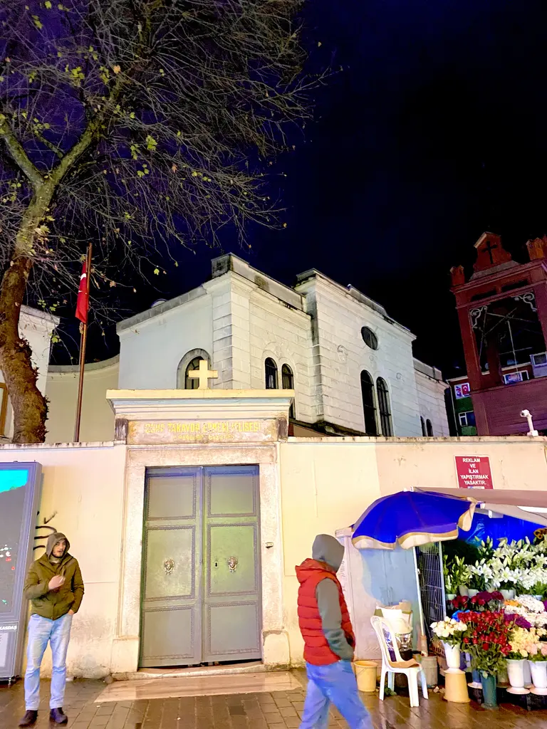 Gece Surp Takavor Kilisesi; aydınlatılmış beyaz cephe, haç, çan kulesi ve önünden geçen insanlar.
