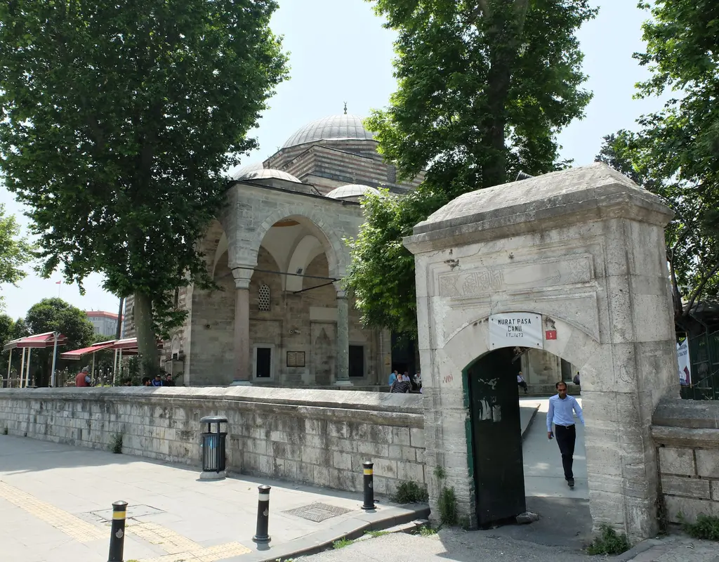 Murat Paşa Camii'nin Millet Caddesi'ne açılan avlu kapısı; taş kapı yapısı, alçak avlu duvarı, çınarlar, arkada revak ve kubbe.