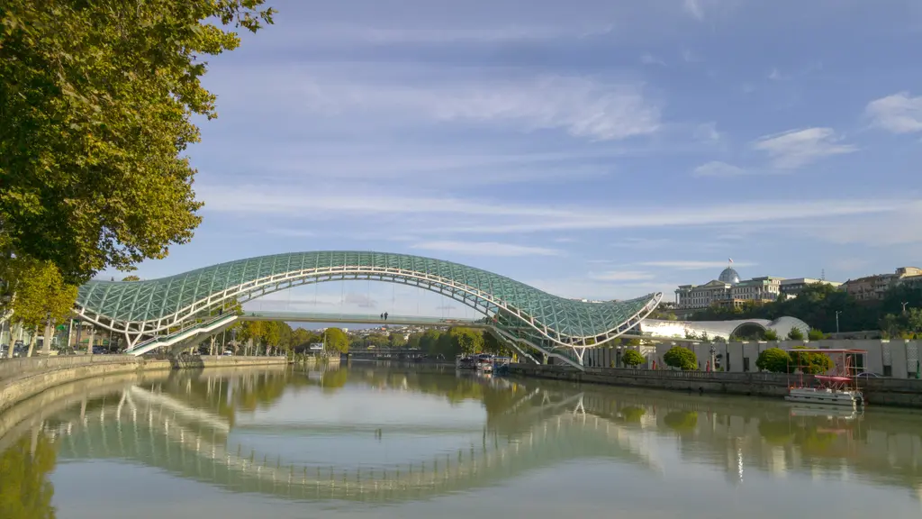 Tiflis'te Mtkvari Nehri üzerinde uzanan Barış Köprüsü; dalgalı cam-çelik çatısı, beyaz taşıyıcı kemerleri ve suya vuran yansıması.