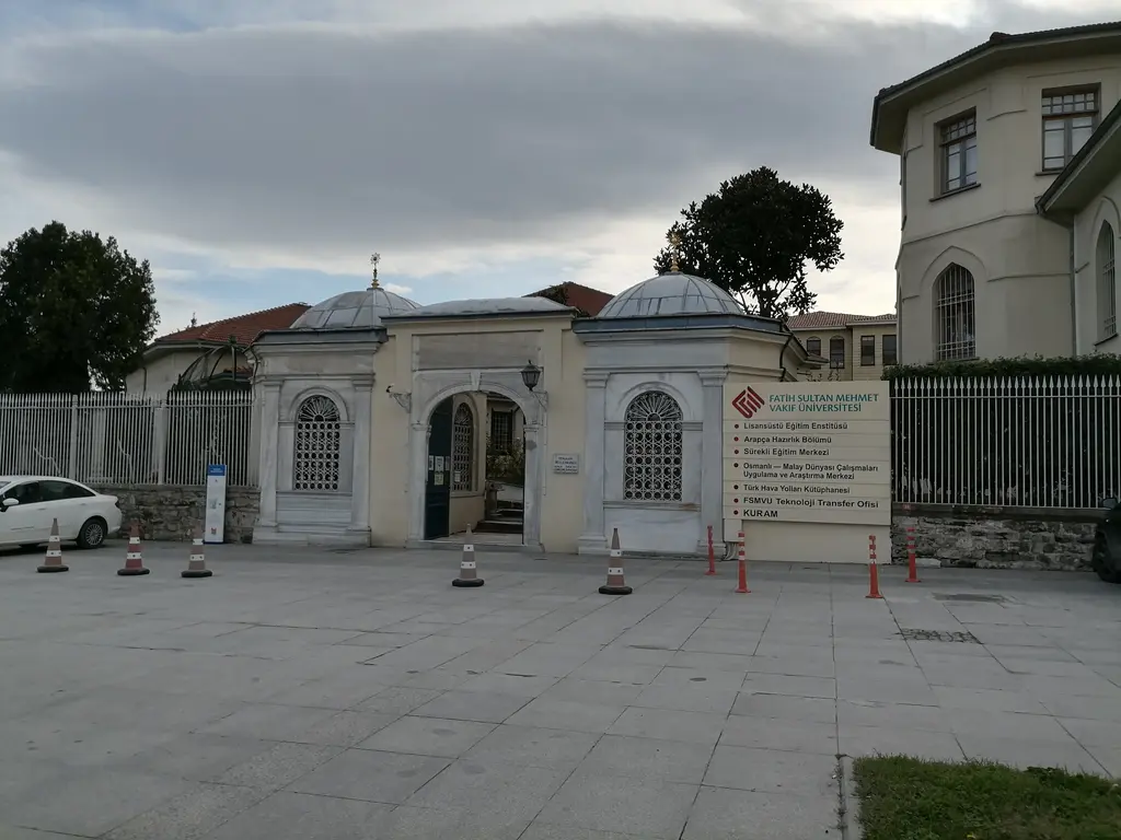Yenikapı Mevlevihanesi'nin girişi; kemerli taş kapı, iki yanında kubbeli küçük yapılar, demir parmaklık ve geniş taş döşeli avlu.