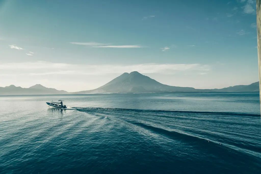 Guatemala'da Atitlán Gölü'nün sabah puslu sakin suları; arkada koni biçimli yanardağlar ve iz bırakarak ilerleyen küçük bir tur teknesi.