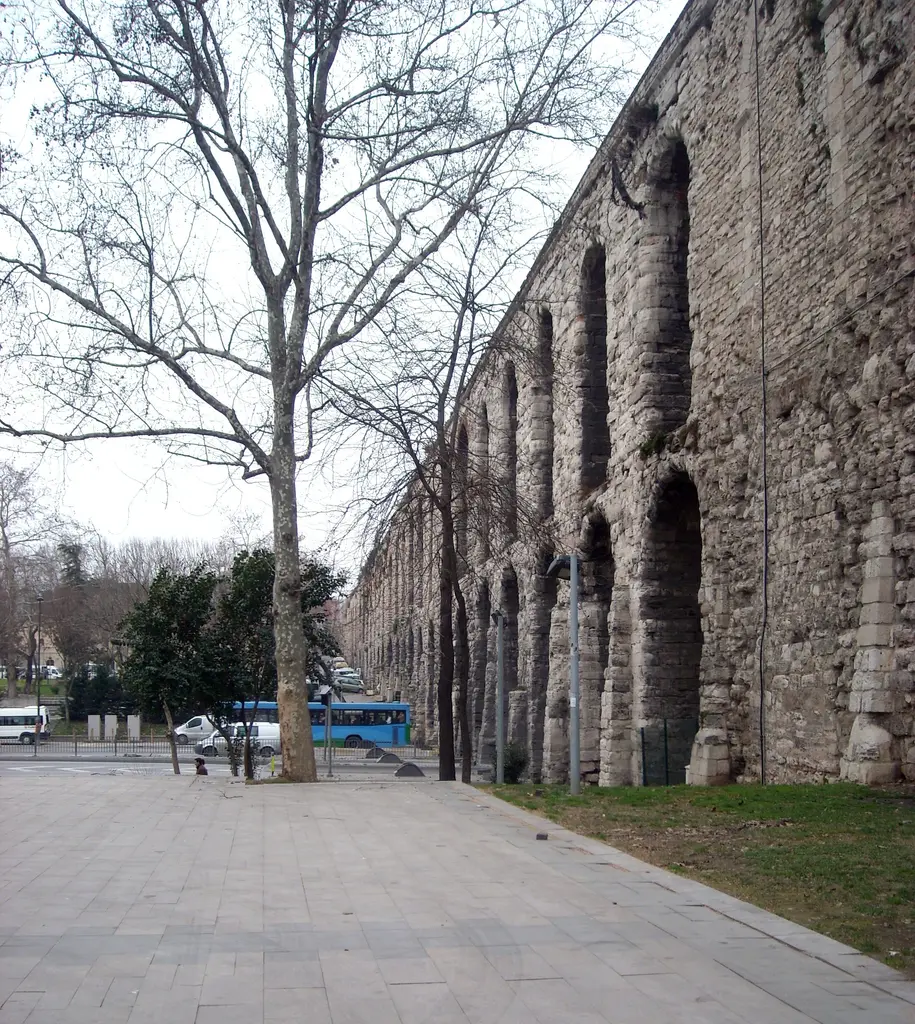 Bozdoğan Kemeri'nin park tarafı; önde geniş gri taş döşeli meydan ve ortasında yapraksız devasa bir çınar, sağda kemer dikey olarak yükseliyor, solda yeşil çim ile ağaçlar.