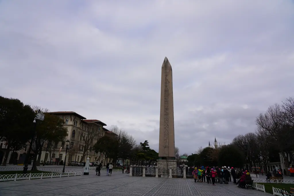 Sultanahmet'te hipodrom meydanındaki Dikilitaş; taş kaide üzerinde yükselen pembe granit sütun, çevresindeki korkuluk ve bulutlu gökyüzü.