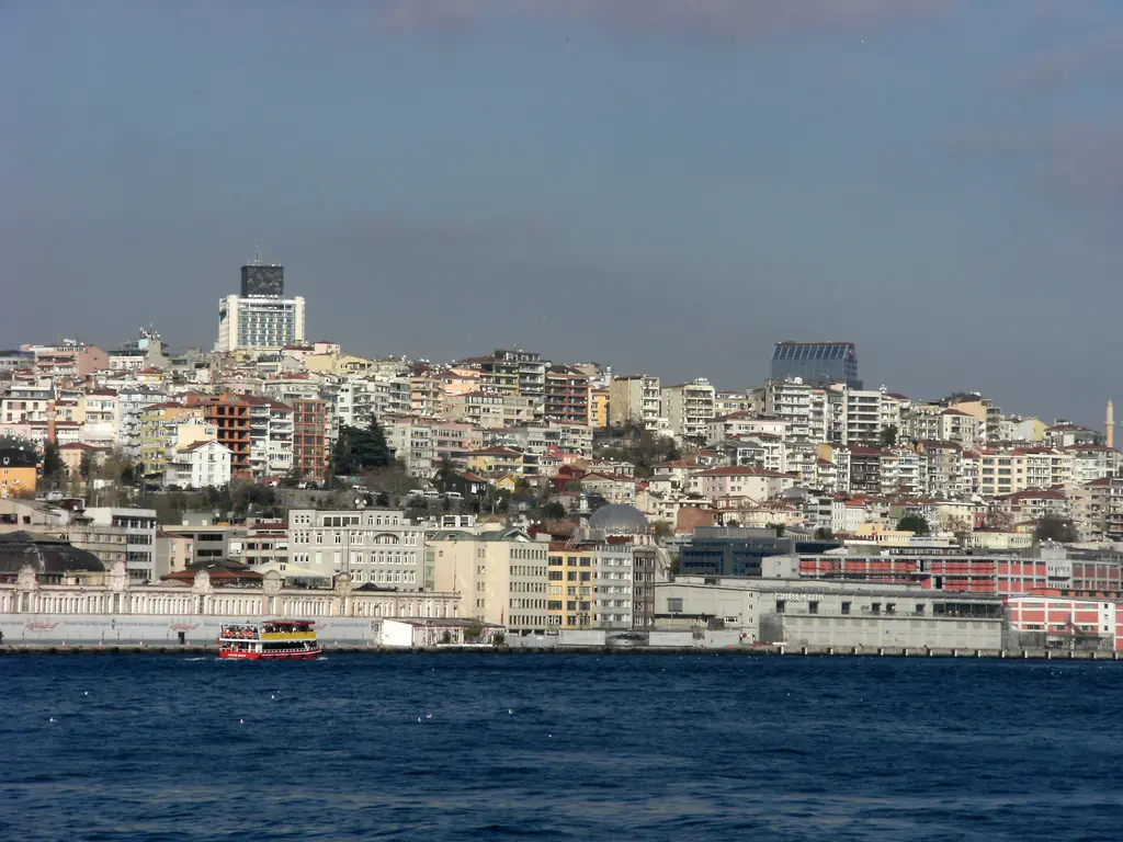 Boğaz'dan Tophane kıyısı; ön planda koyu mavi deniz, kıyıda alçak yapılar, yamaçta sık apartmanlar ve tepede yüksek bir bina.