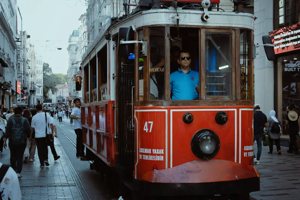 İstiklal Caddesi'nde kalabalığın ortasından geçen kırmızı nostaljik tramvay; iki yanda eski taş cepheli binalar ve rayların üzerinde yürüyen insanlar.