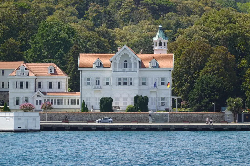 Alman Büyükelçiliği'nin Tarabya yazlığı; kıyıda beyaz köşk, kuleli çatı, bayraklar ve arkada ağaçlık park.