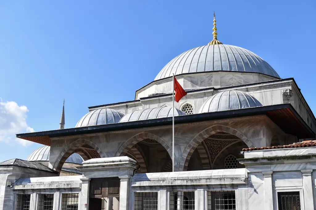 Sultanahmet'te Sultan Ahmed Türbesi; mermer kaplı gövde, dilimli kurşun kubbe, önde kemerli revak, yanında küçük kubbeler ve direkte Türk bayrağı.