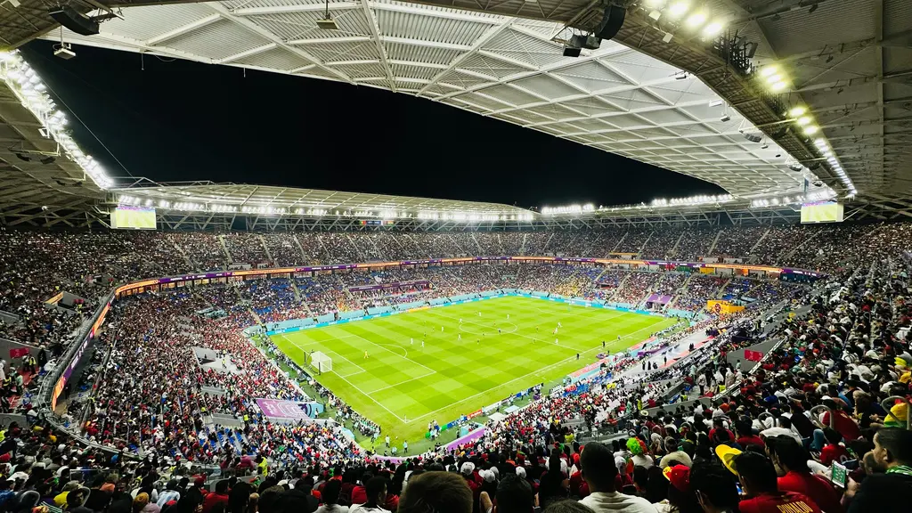 Doha'da tıklım tıklım bir stadyum; yeşil sahayı çevreleyen dolu tribünler, üstteki açık çatı boşluğu ve kenardaki aydınlatma sıraları.