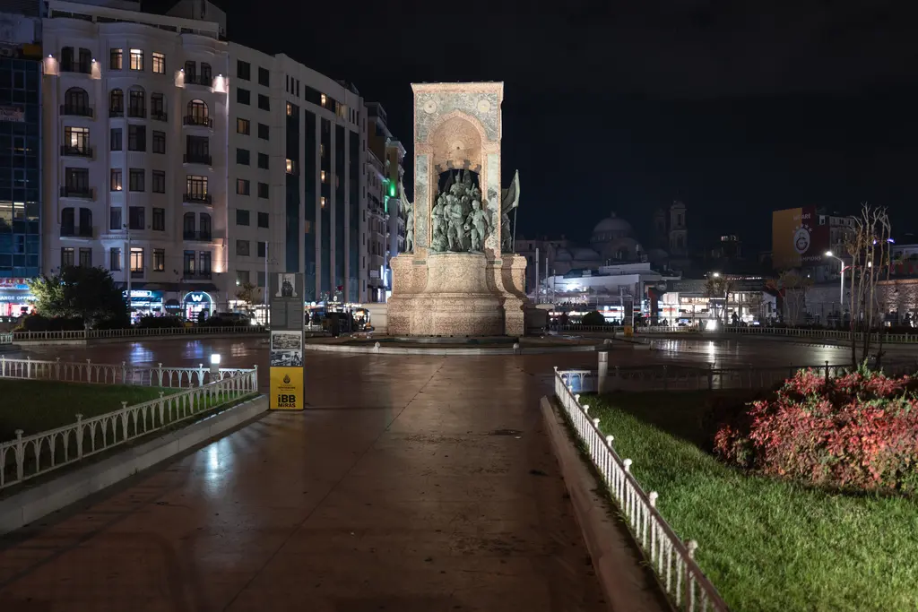 Gece Taksim Meydanı'nda ışıklandırılmış Cumhuriyet Anıtı; önünde ıslak taş yol, iki yanda çiçekli bahçeler ve arkada aydınlık binalar.