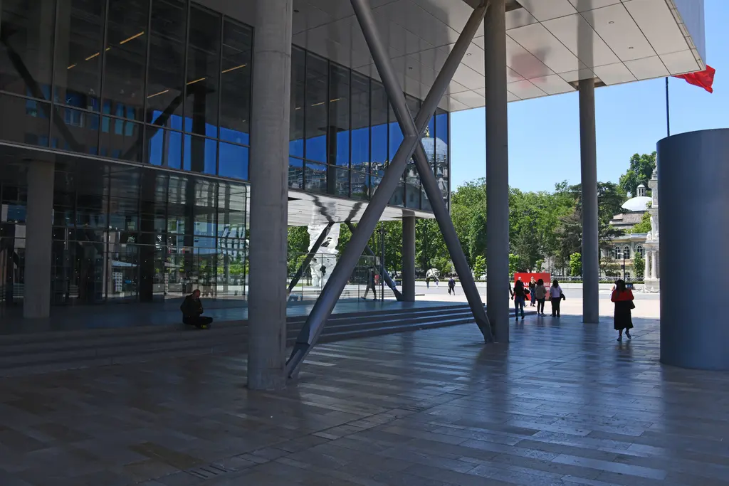 İstanbul Modern'in saçağı altında gölgeli alan; ince beyaz sütunlar, çapraz çelik gergiler ve ileride güneşli meydan.