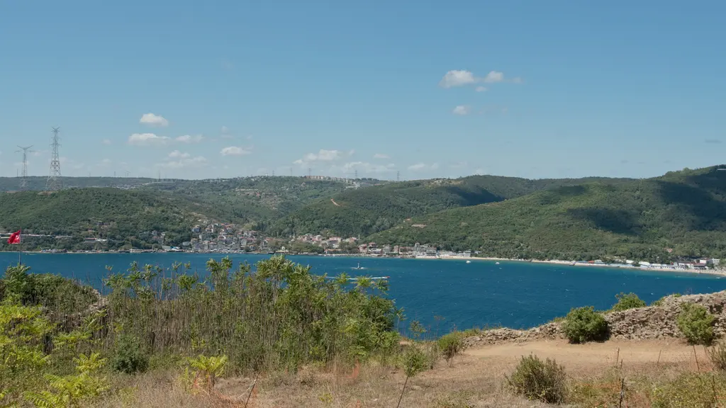 Yoros Kalesi'nden Boğaz; kuru otlu yamaçtan görünen lacivert deniz, karşı kıyıda köy ve ormanlık tepeler.