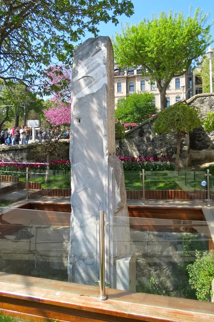 İlkbaharda Milyon Taşı; cam korkuluk içinde mermer ayak, arkada pembe çiçekli ağaçlar, lale tarhları ve kaldırımda yürüyen insanlar.
