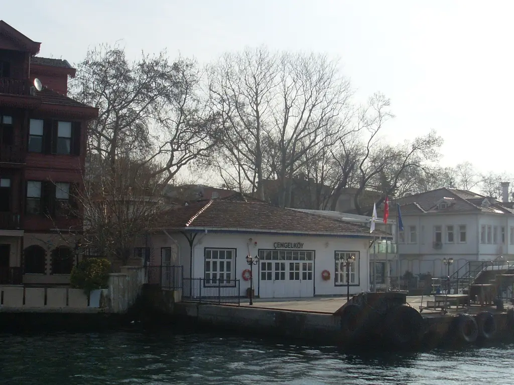 Çengelköy İskelesi'ne denizden bakış; beyaz ahşap iskele binası, yanında kırmızı ev ve arkada yapraksız ağaçlar.